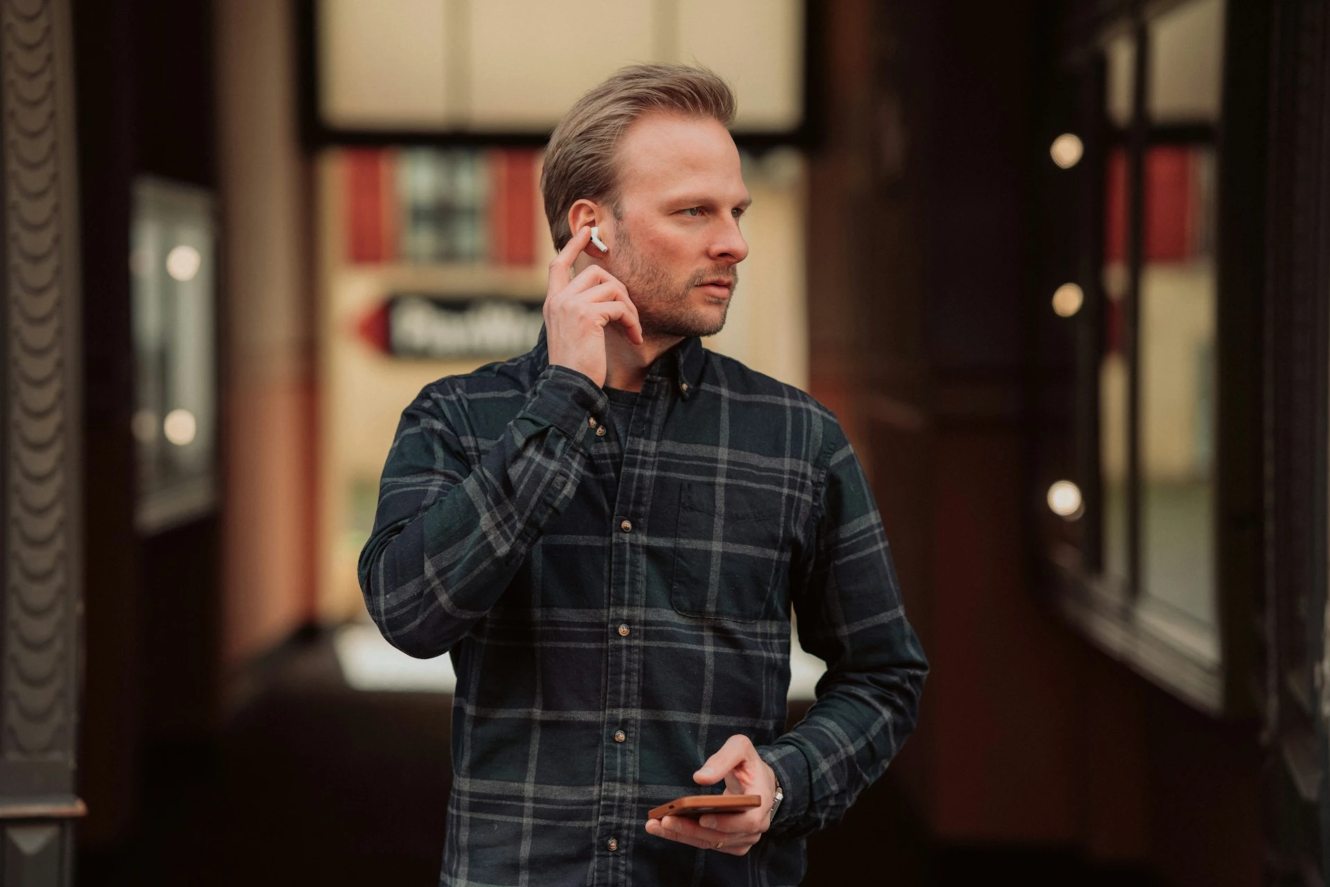 A man in a plaid shirt with an earbud in his ear, holding a phone.