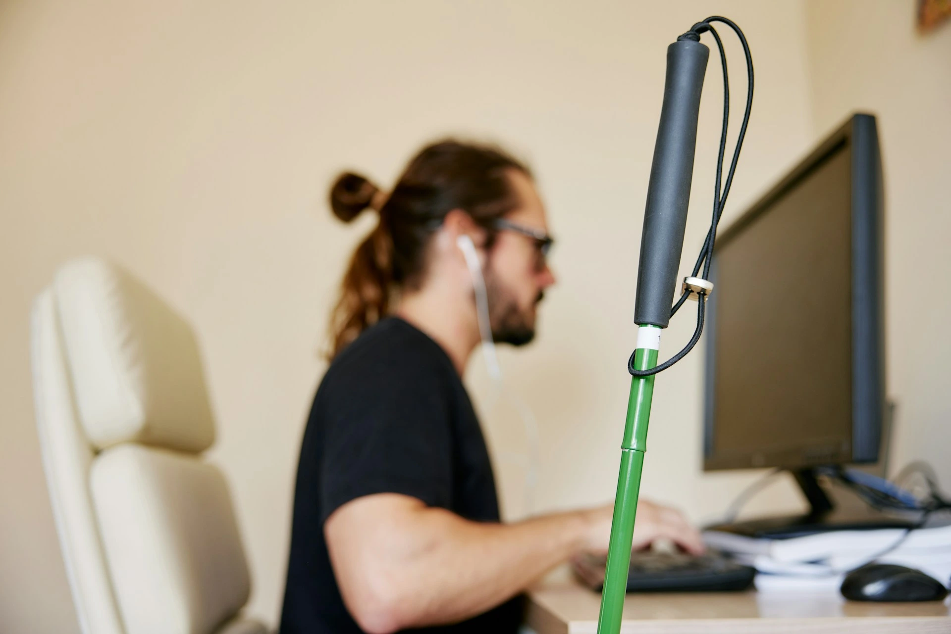 Blurred profile of a man at a computer with a green white-cane in sharp focus in the foreground.