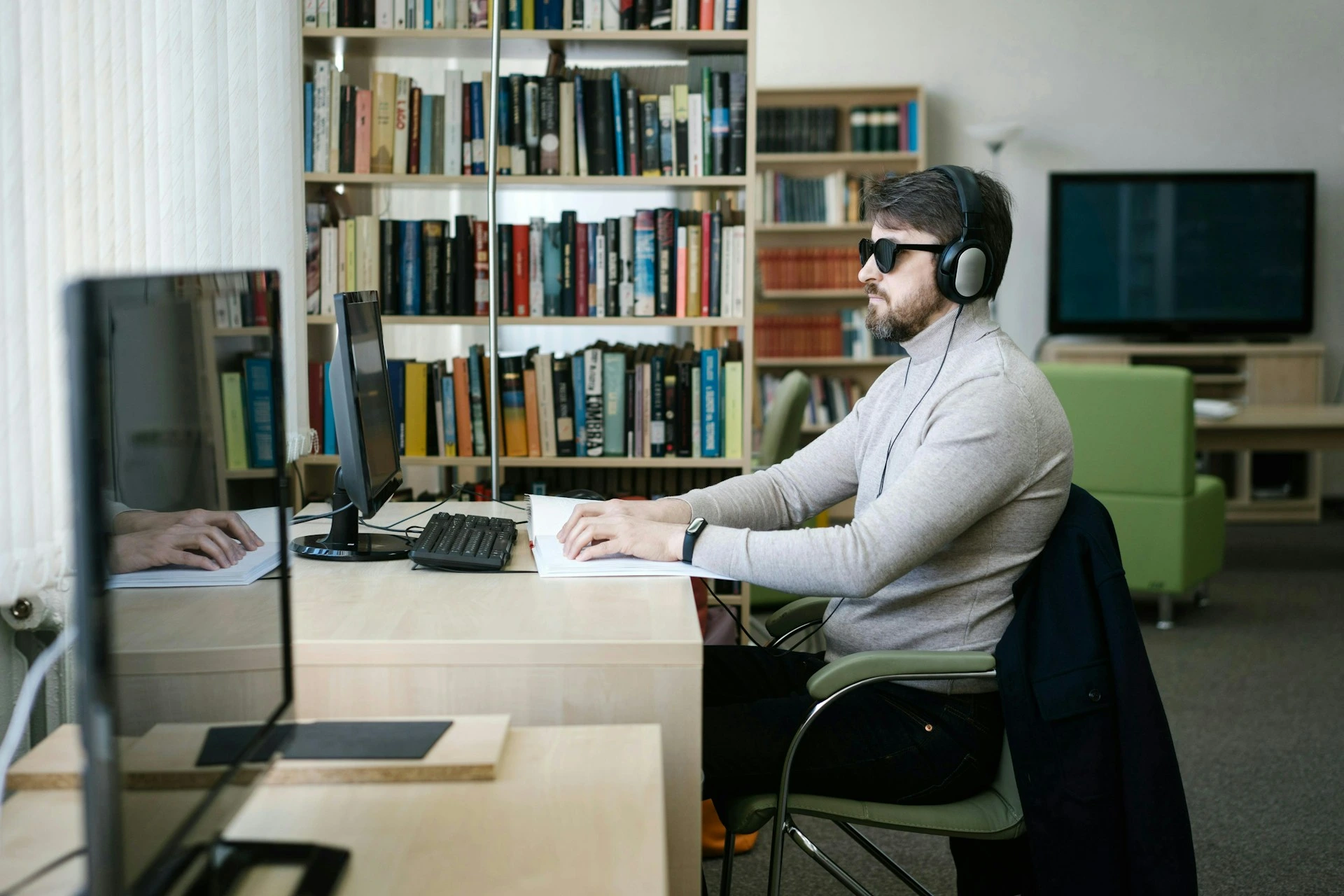 A man wearing dark sunglasses and headphones sitting at a desk with a computer and braille book.