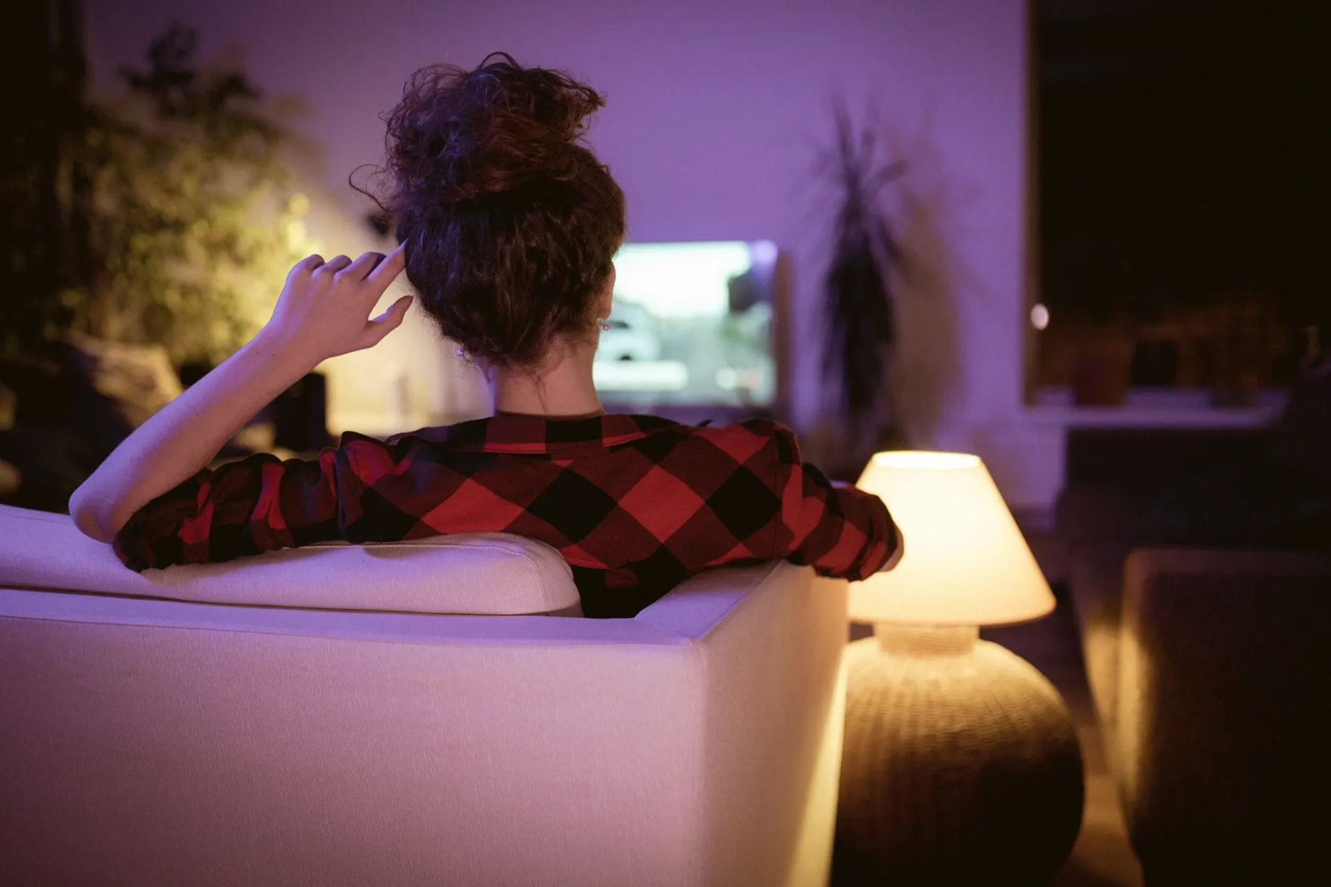 A person sits on a couch watching a TV in a dimly lit room.