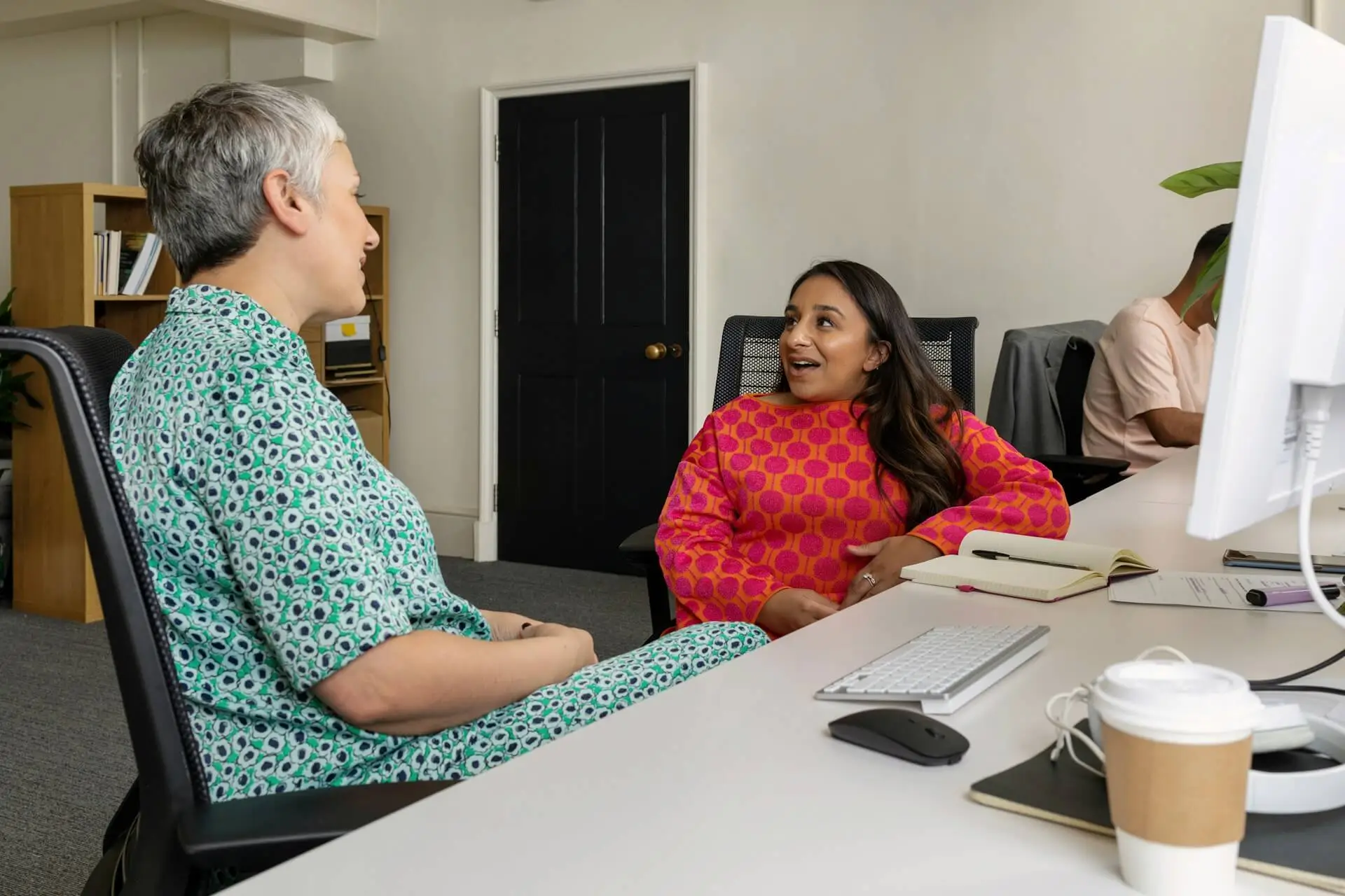 Two women engage in a friendly conversation at a white desk in an office setting.