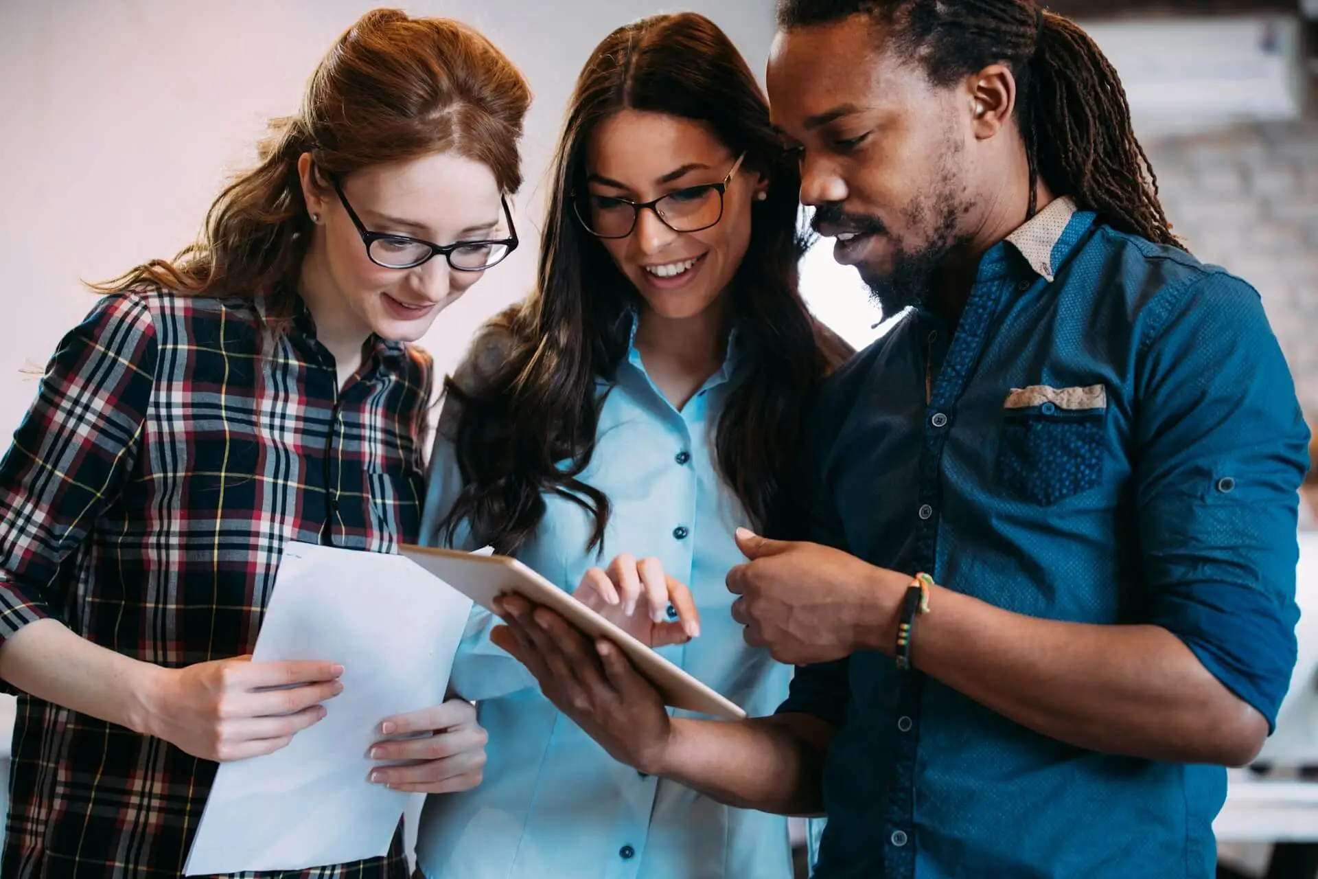 Three colleagues look together at a tablet screen while smiling in a bright office.