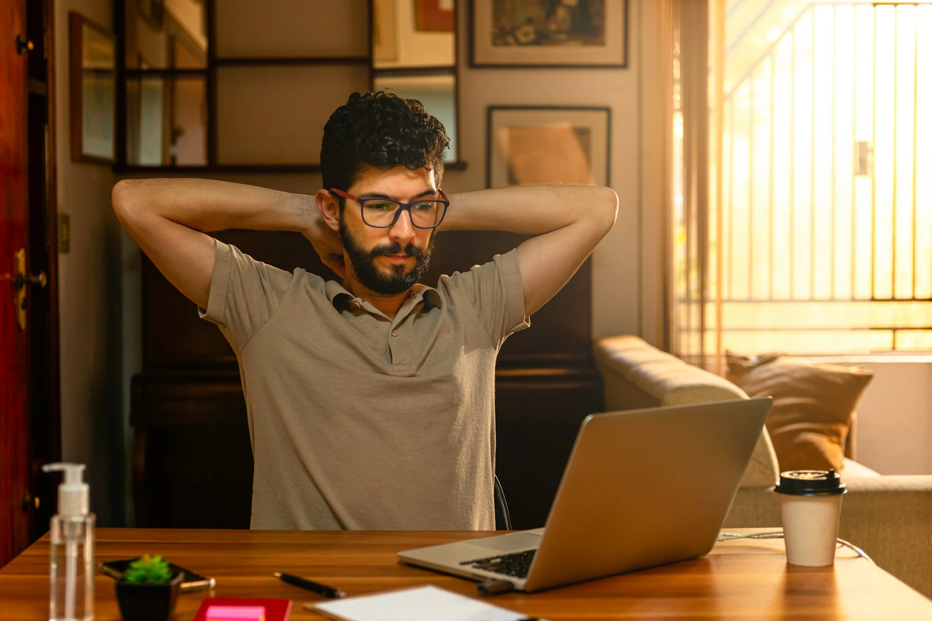 A man with glasses sitting at a desk, looking at his laptop with his hands behind his head.