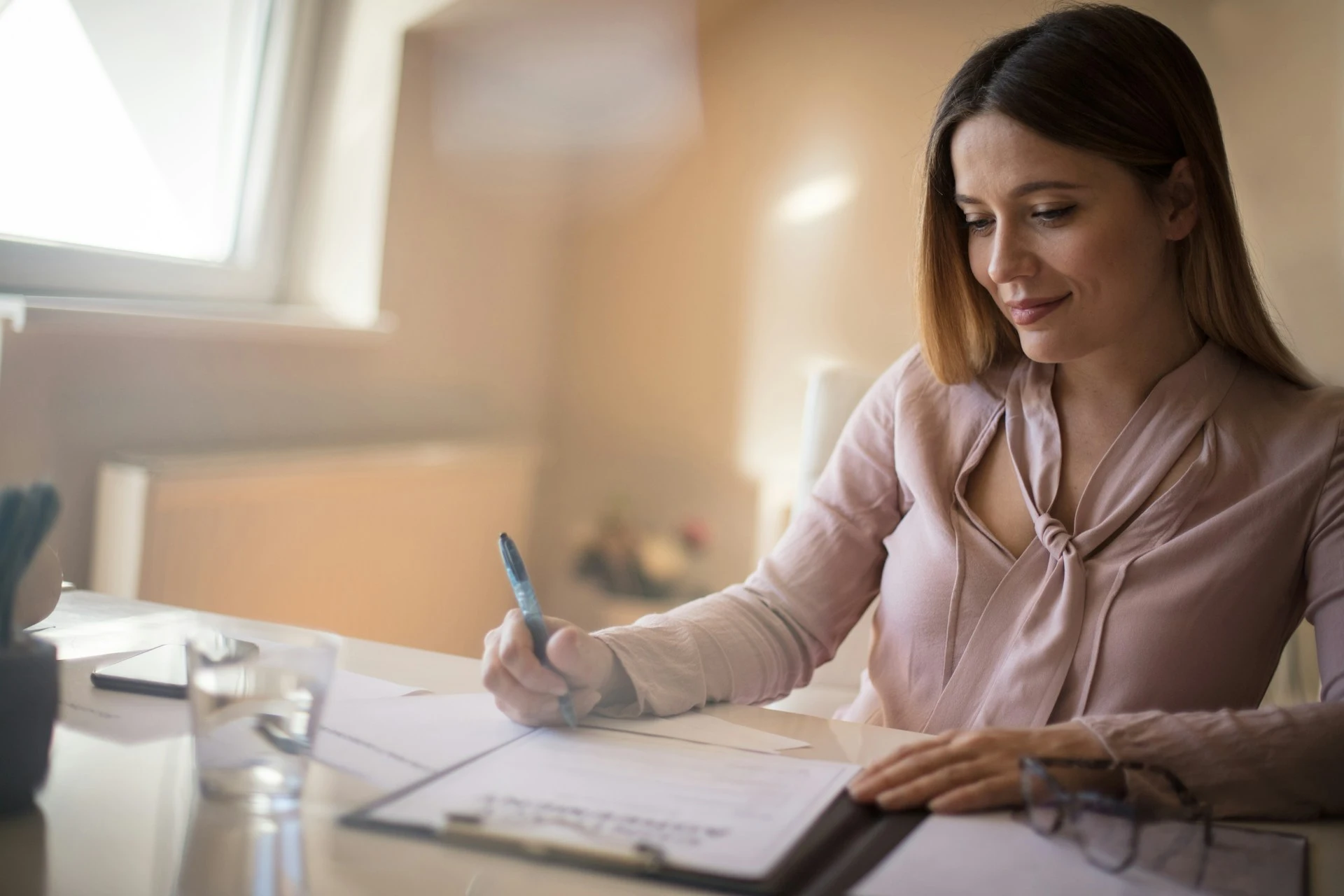 A woman sitting at a desk and writing on a document held in a clipboard.