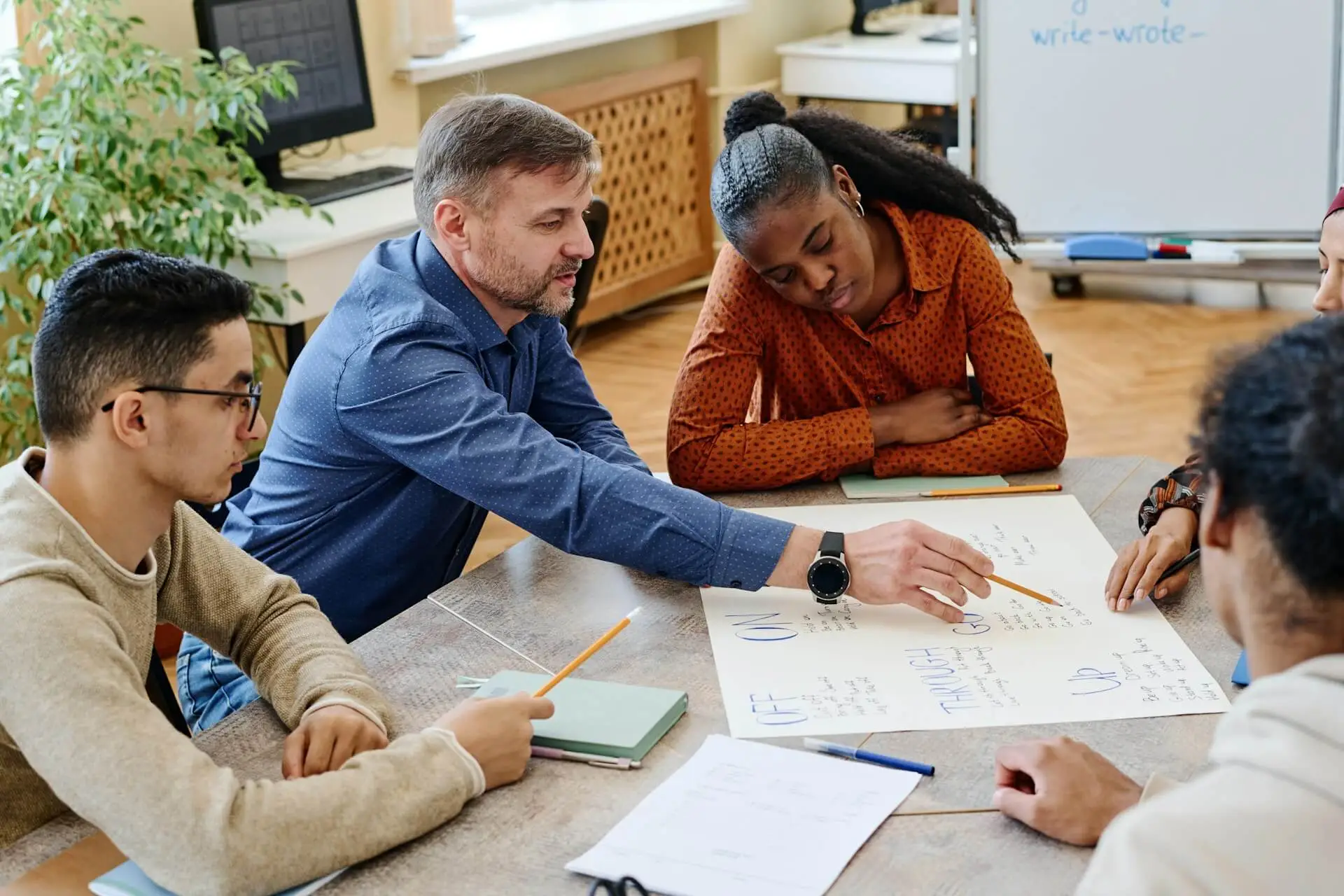 A diverse group of students sits around a table while an instructor points to a large sheet of paper.