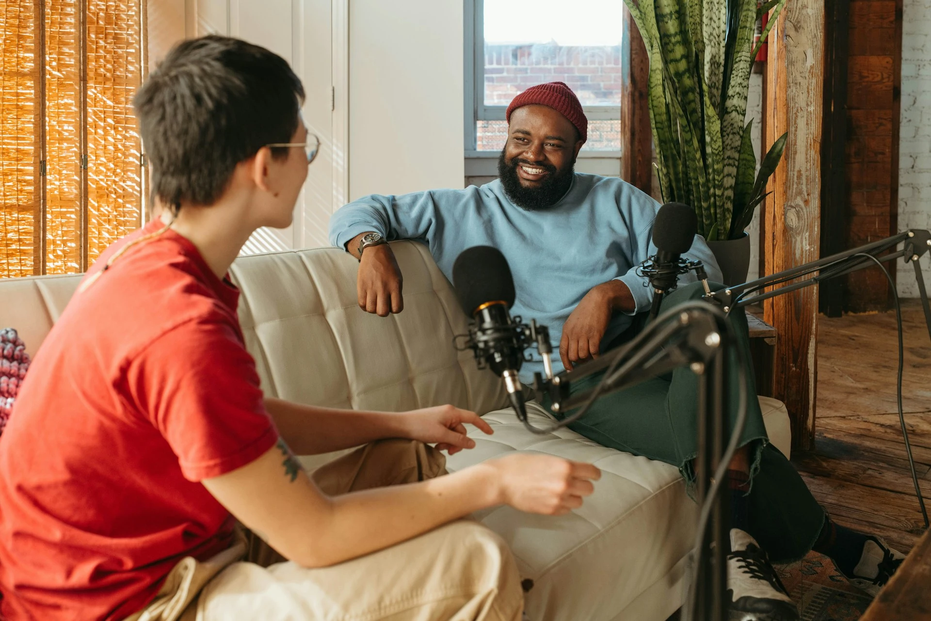 Two people sitting on a couch facing each other, speaking into microphones on boom arms.