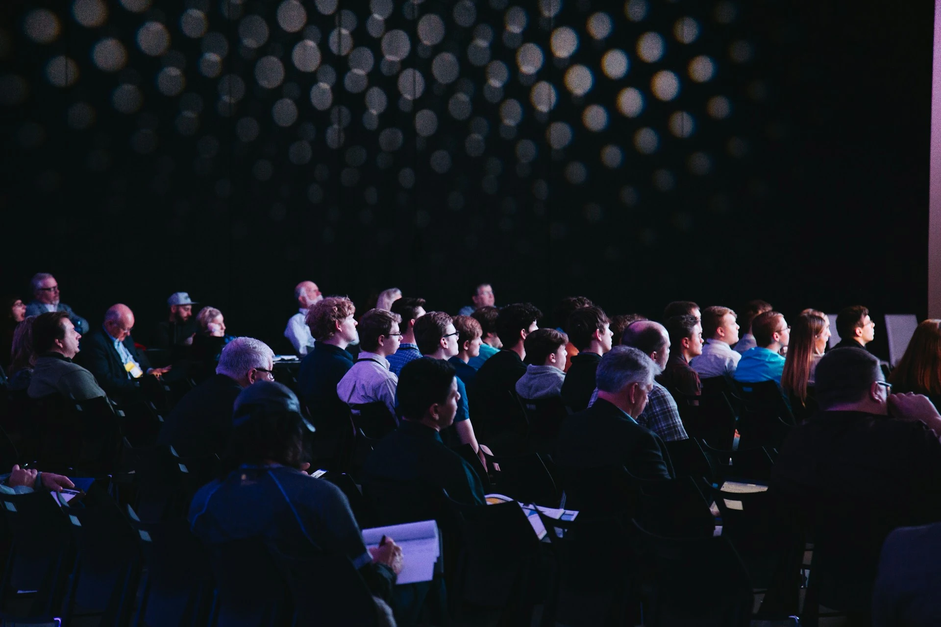 A side view of an audience sitting in a dark auditorium, looking forward at a stage.
