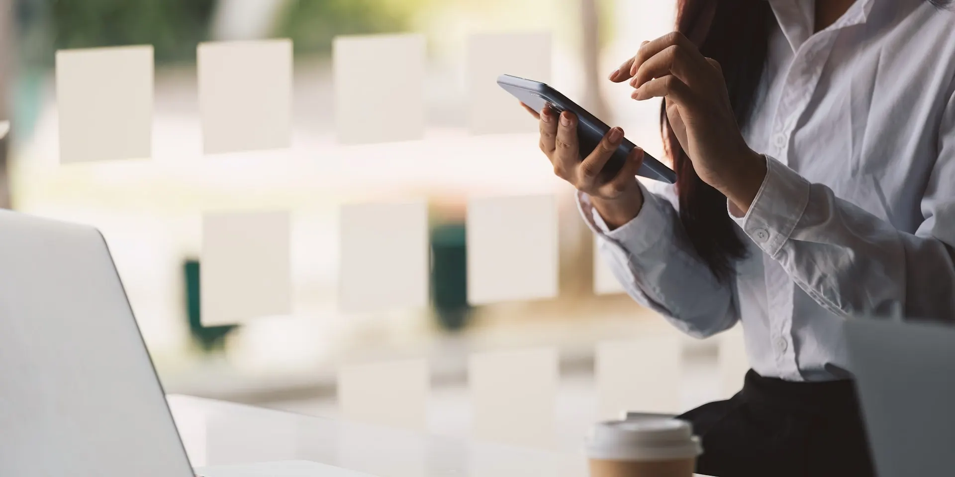 A close-up of a person sitting at a desk using a smartphone, with a laptop in the background.