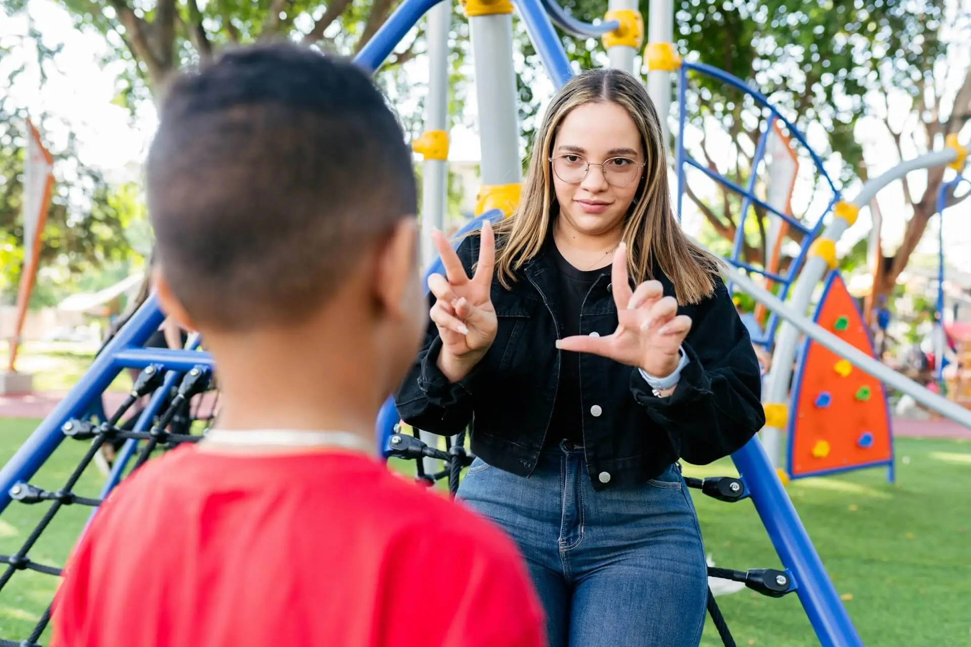 A woman uses sign language to communicate with a young boy at an outdoor playground.