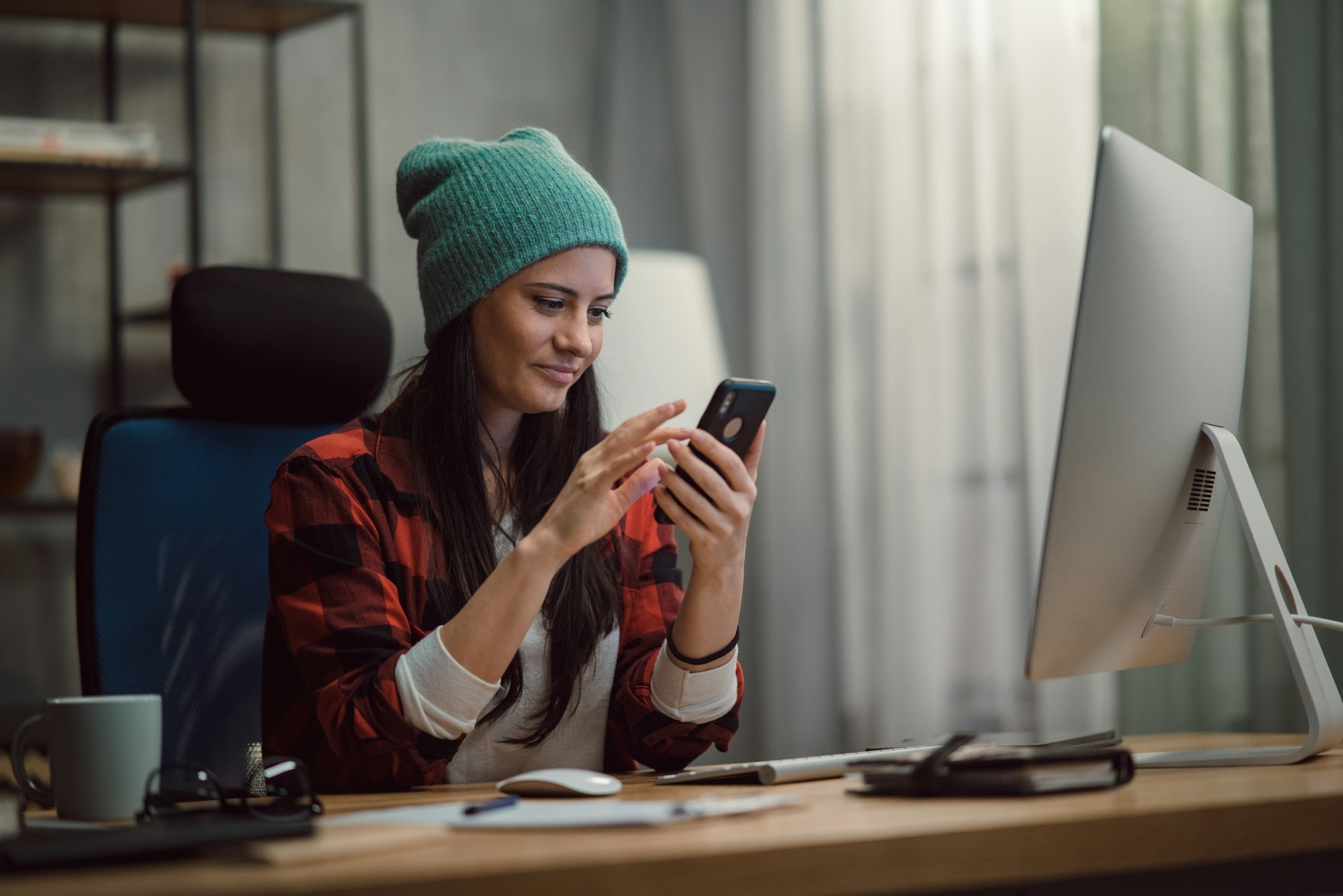Woman in a beanie and plaid shirt using a phone at a desk.