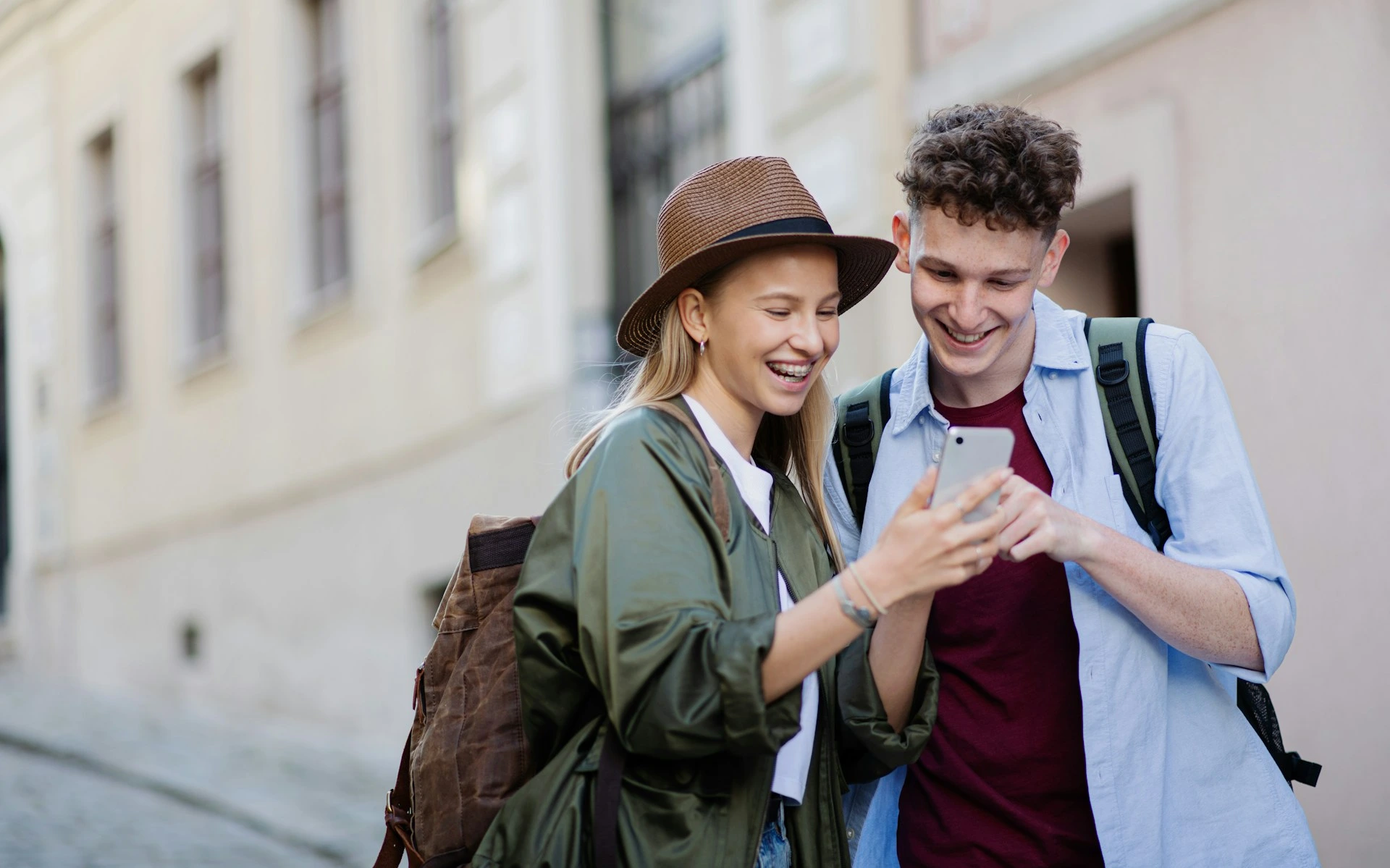 Two young travelers with backpacks smiling while looking at a smartphone on a city street.