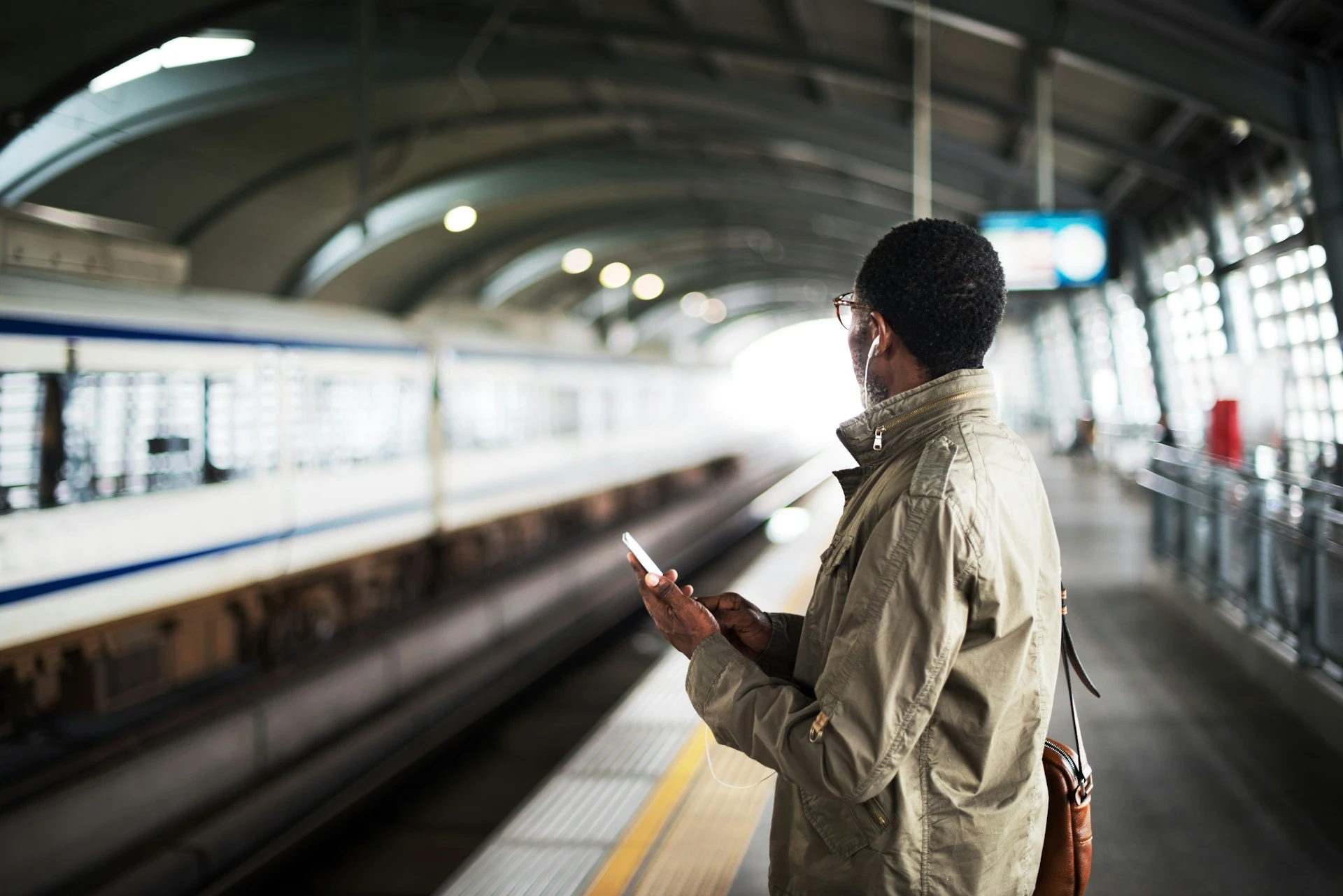  A man stands on a train platform, holding a phone and wearing headphones.