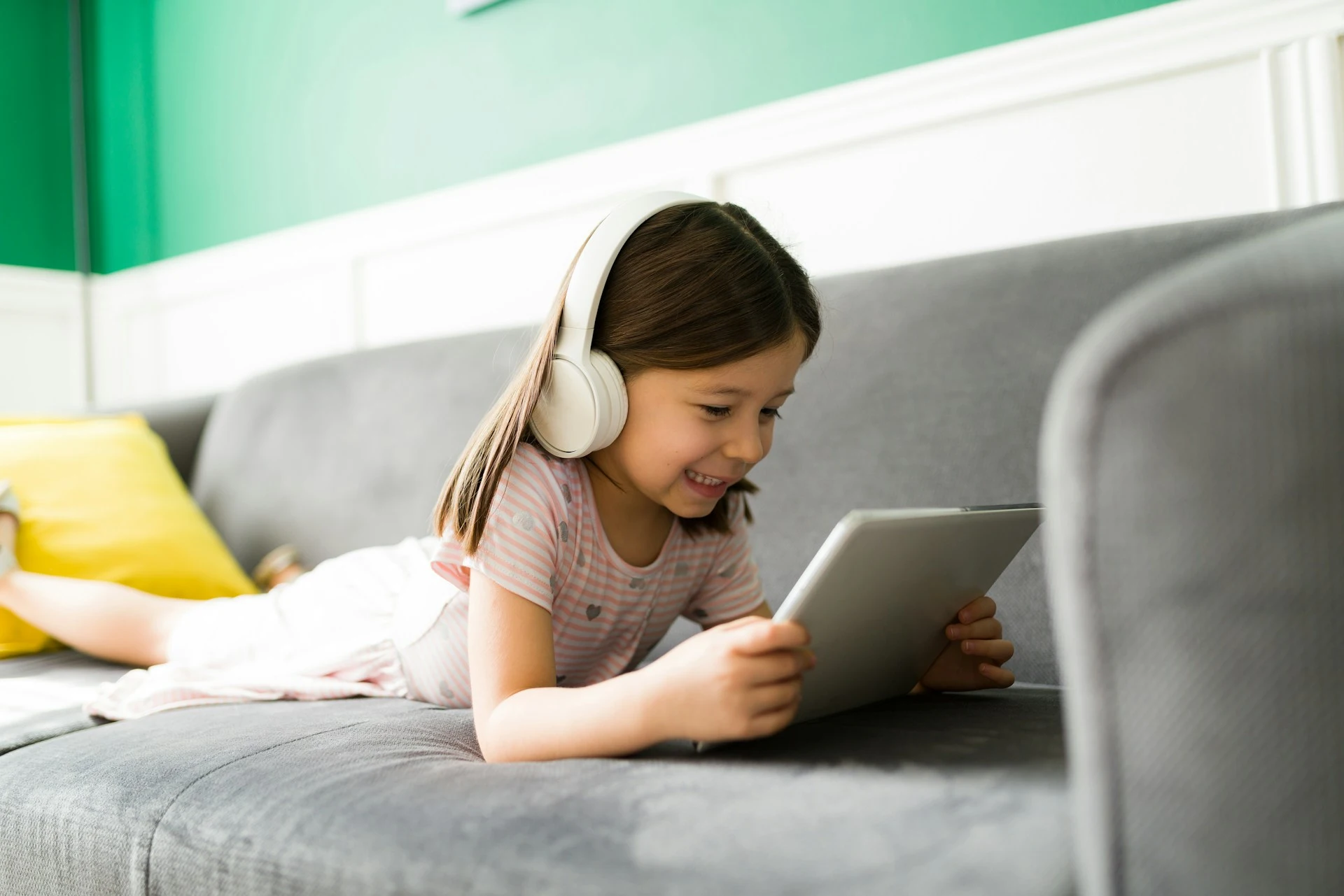 A young girl lying on a couch wears white headphones and smiles while looking at a tablet.
