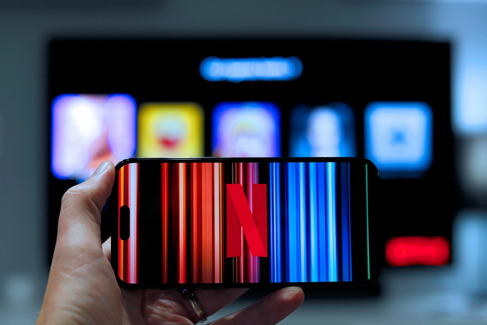  A hand holds a smartphone displaying the Netflix logo, while a TV screen showing blurred content is visible in the background.