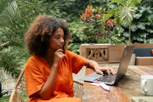 A woman with curly hair sits outdoors, looking at a laptop with her hand on her chin.
