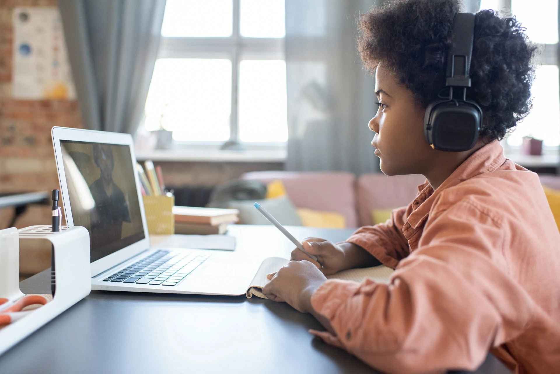 A child with headphones looks at a laptop while holding a pen and notebook.