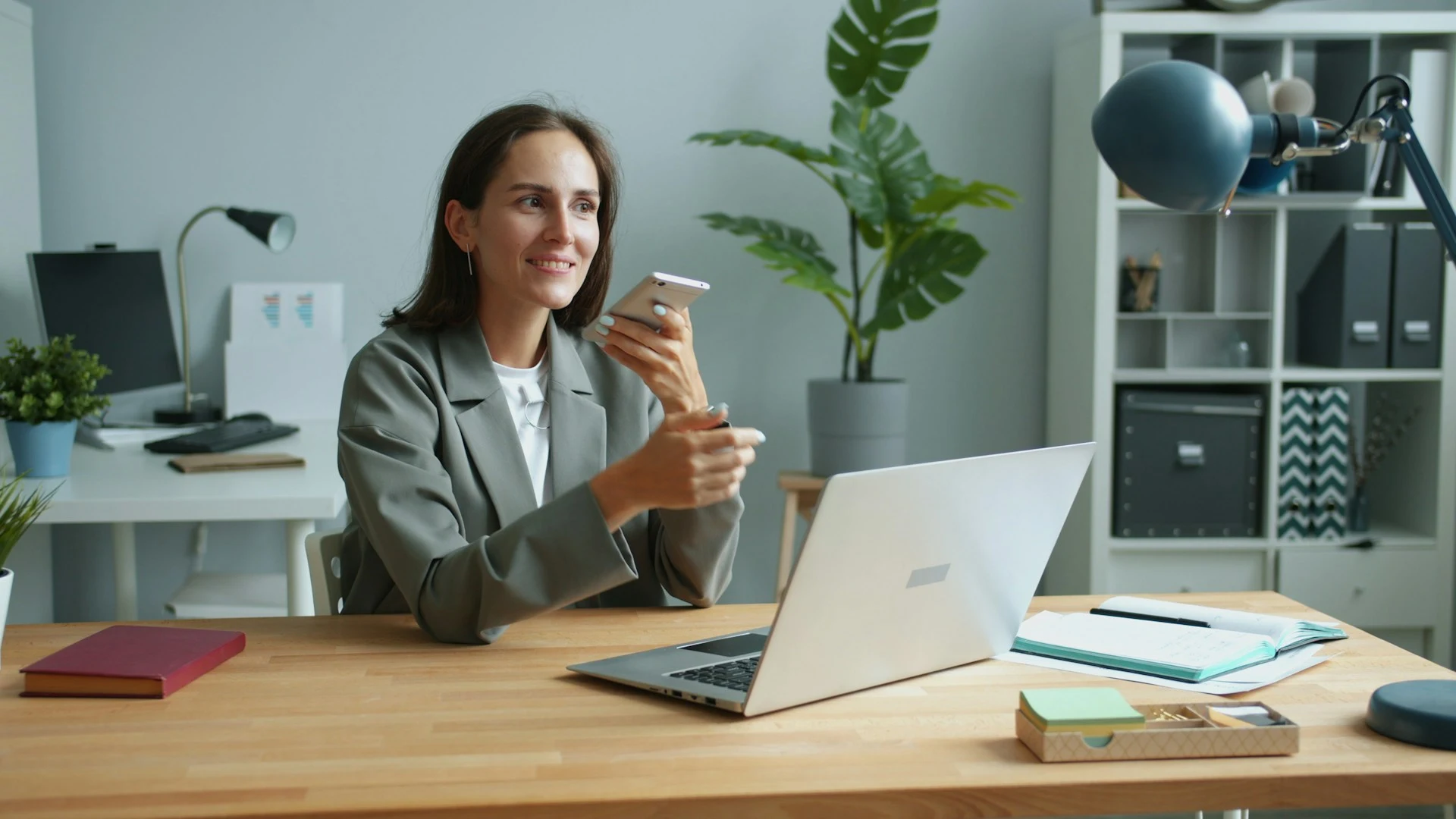 A woman sitting at an office desk with a laptop, holding her smartphone up to record a voice memo.