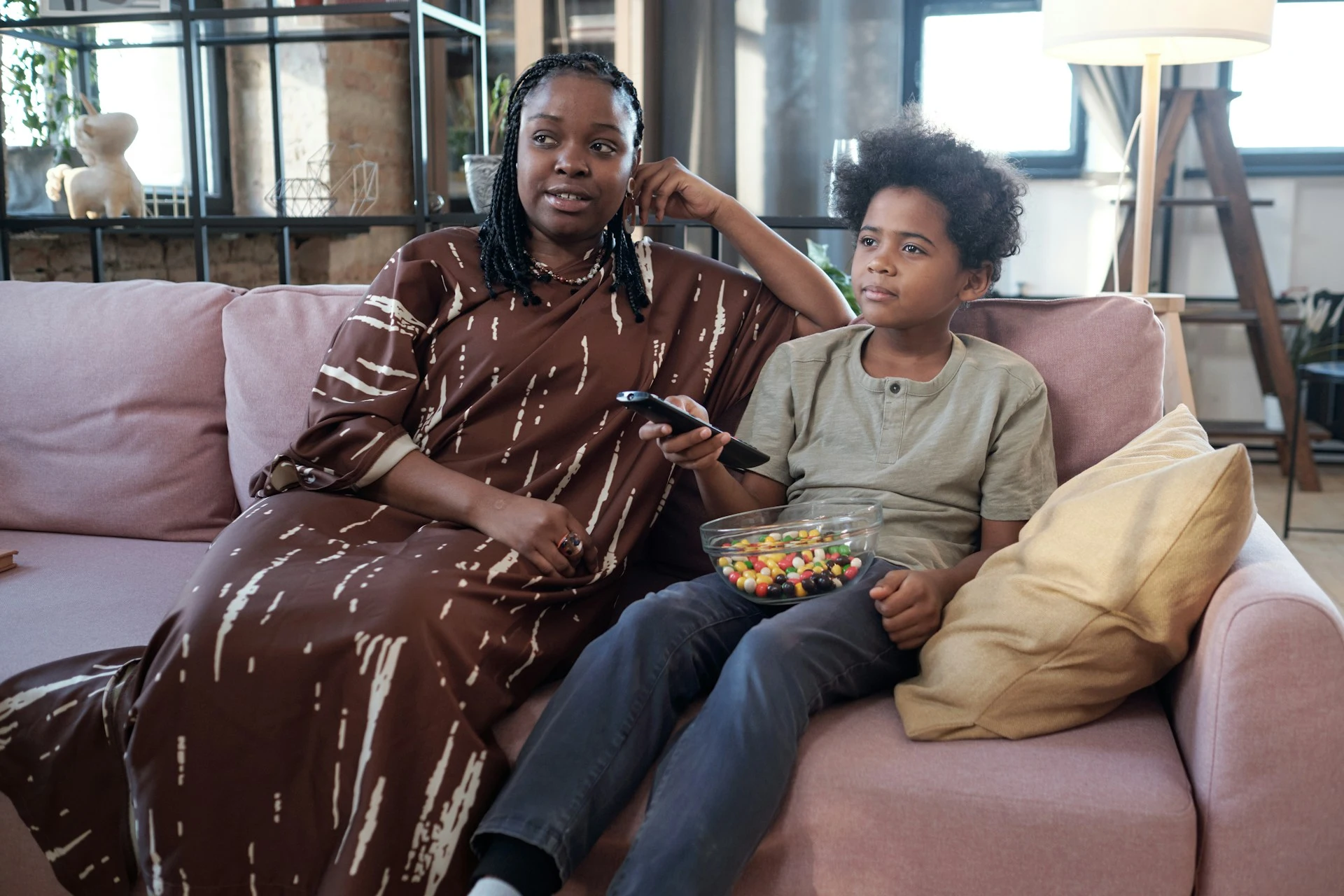 A woman and a boy sitting on a couch, watching TV with a remote and a bowl of candy.