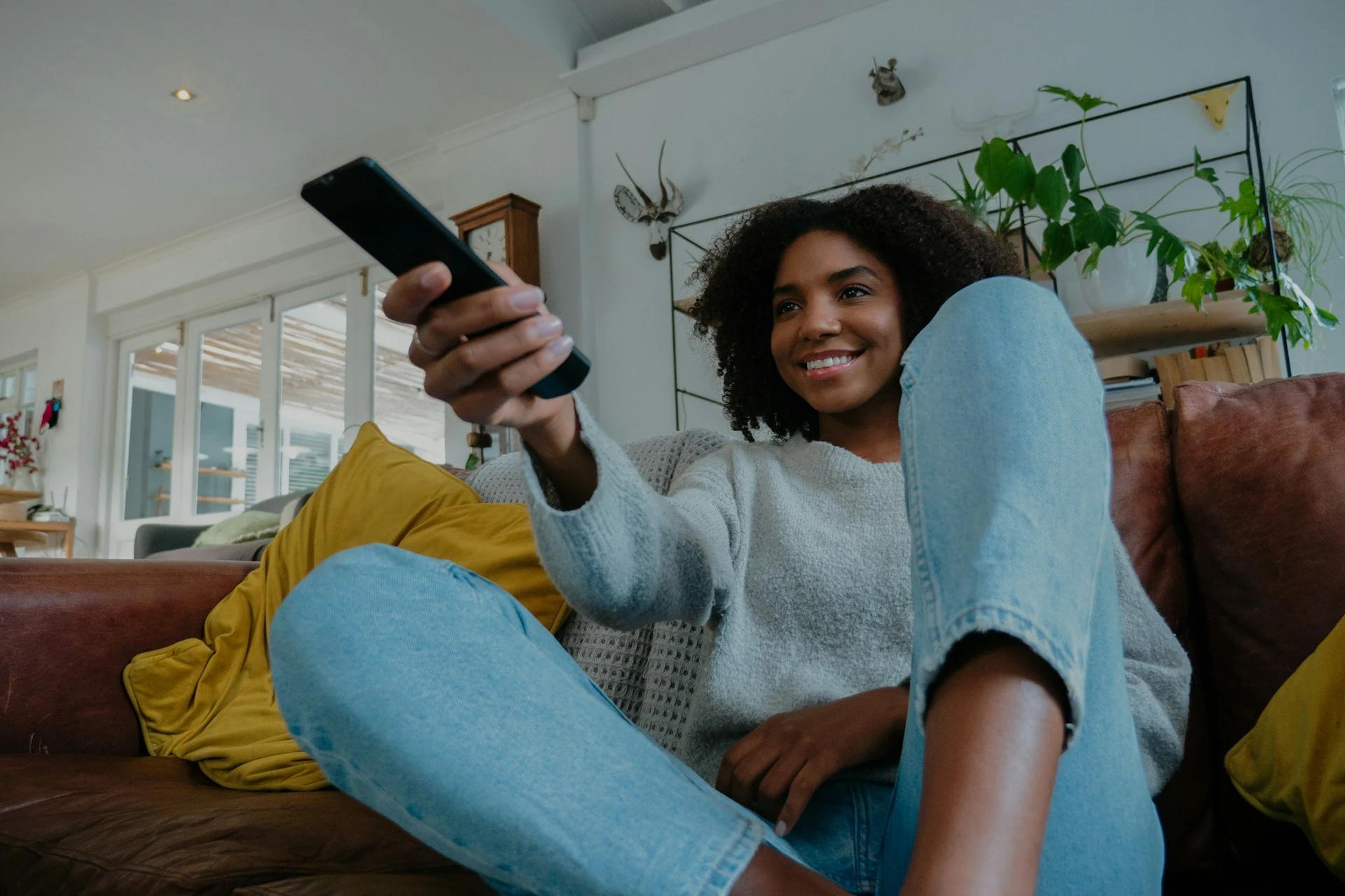 A smiling woman sitting on a couch and holding up a TV remote control.