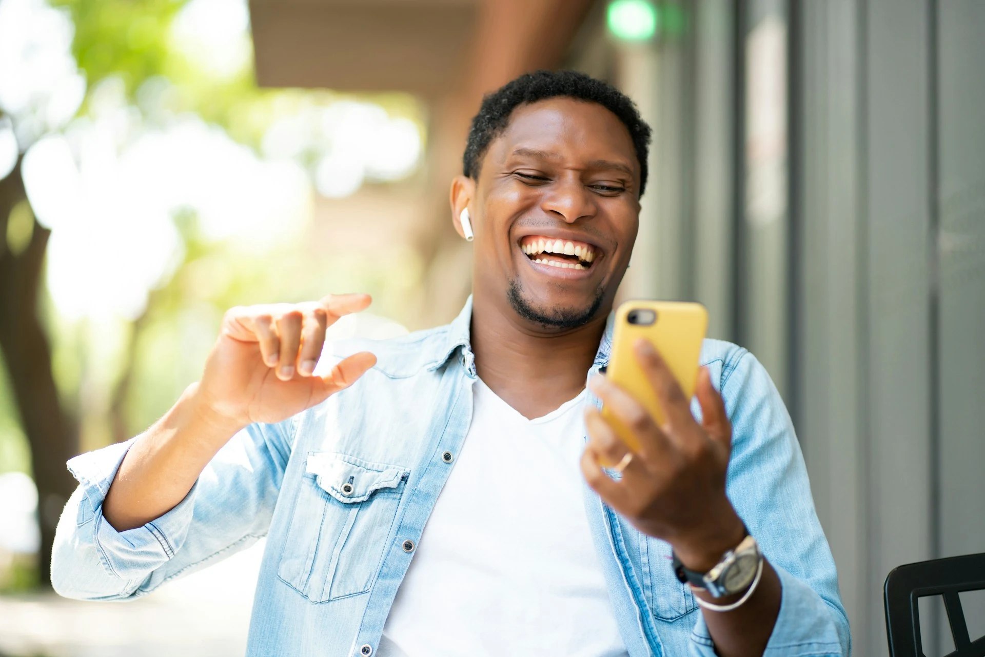 A smiling man wearing earbuds outdoors, laughing while looking at his smartphone.