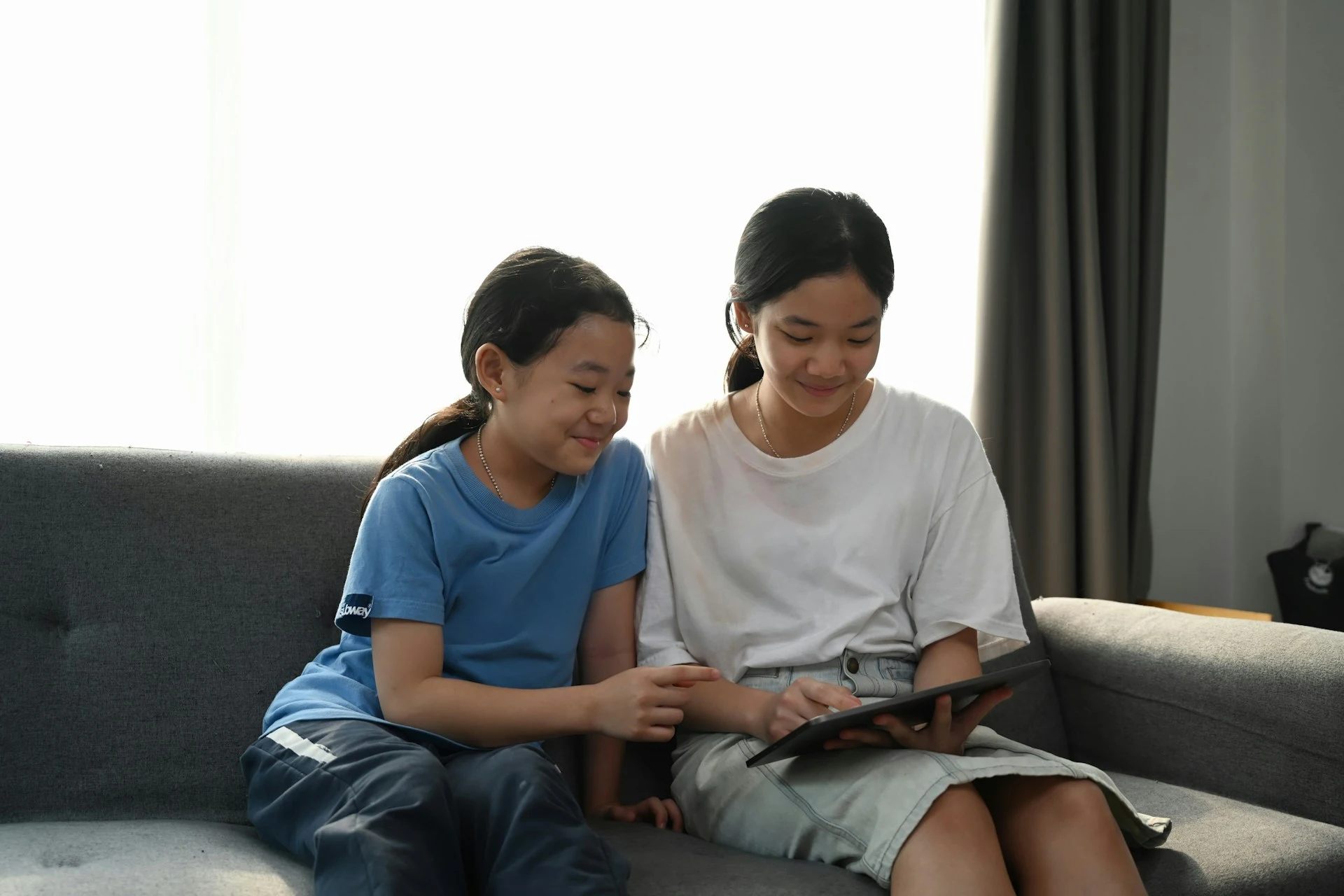 Two young girls sit together on a sofa, smiling as they look at a digital tablet.