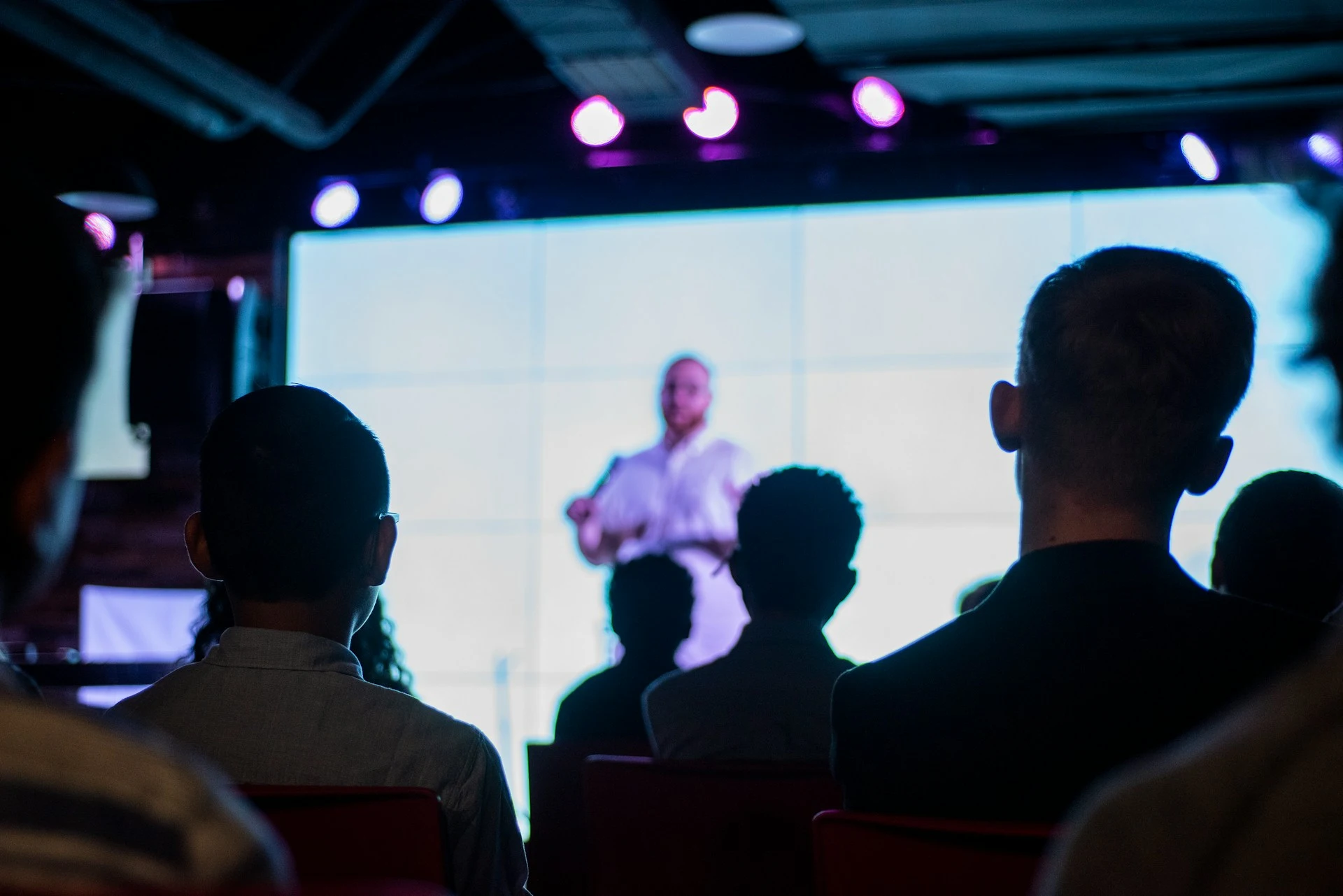 An audience seated in a darkened auditorium watching a speaker give a presentation on stage.