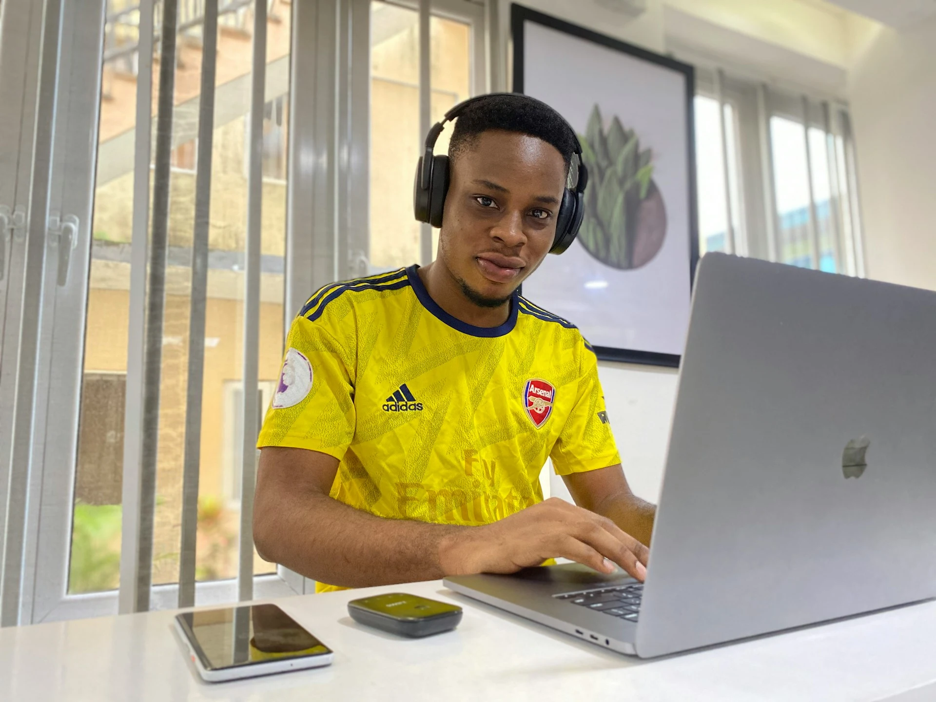 Young man wearing headphones and a yellow Arsenal football jersey sitting at a desk using a laptop.
