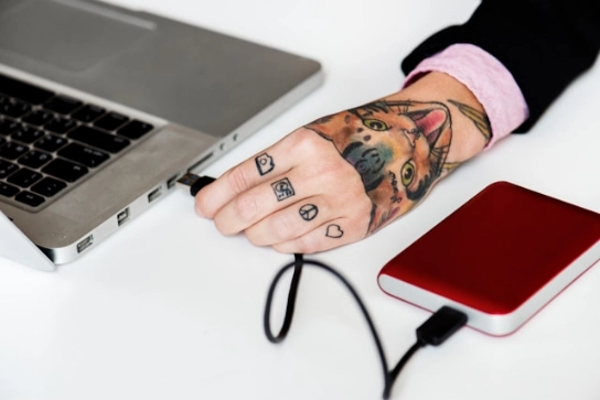 A tattooed hand connecting an external hard drive to the laptop.