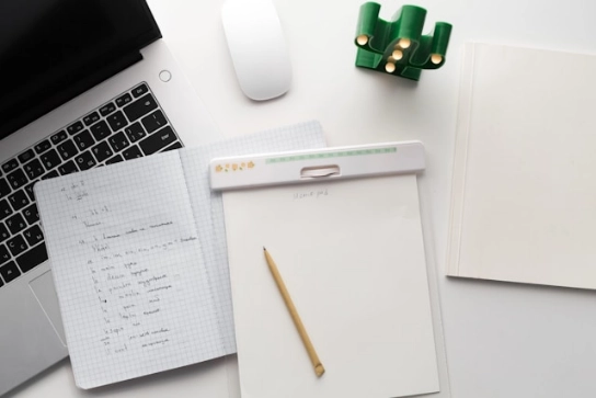 Closeup of of notebooks, a laptop, and a mouse on a white desk.