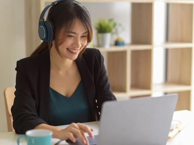 A woman wearing headphones and smiling while working on a laptop.