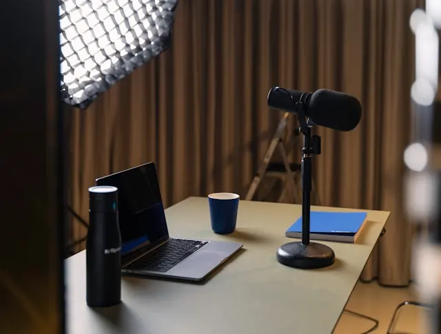 A laptop, notebook, cup, microphone and tumbler on a desk.