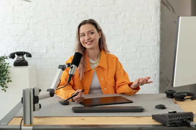 A woman with an orange jacket sitting on a desk and speaking to a mic.