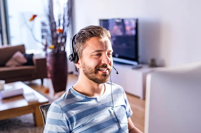 A man with a headphone and a built-in microphone speaking in front of a computer screen.