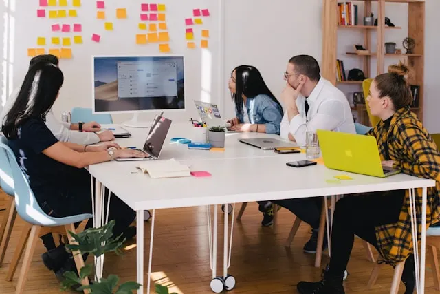 Five person around a table watching a turned on iMac screen.