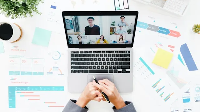 Laptop showing a virtual meeting and related meeting documents on a table.