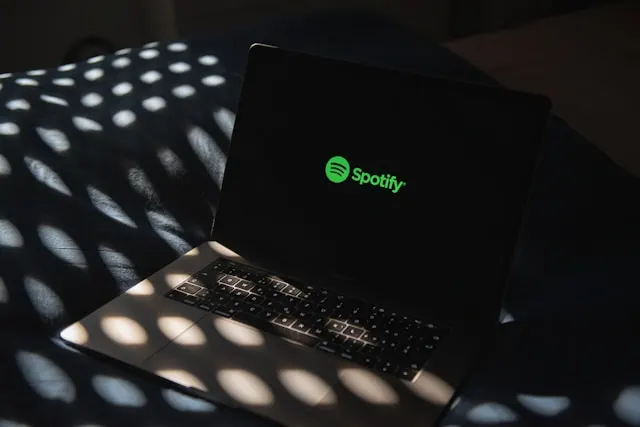 A laptop sitting on top of a bed, its screen showing the Spotify logo.