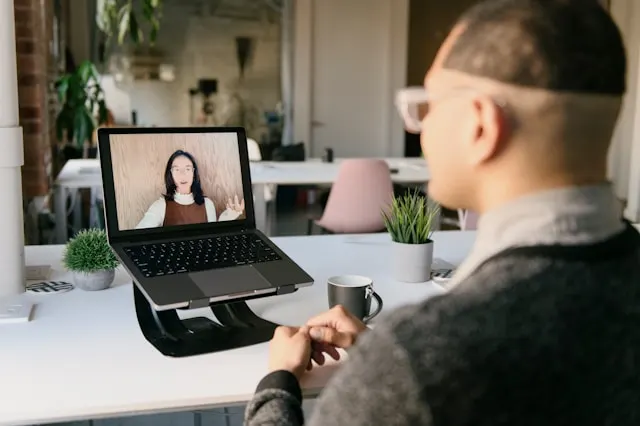 A man watching a woman giving a virtual presentation from his laptop.