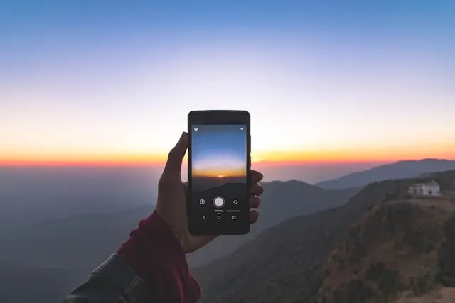 A hand holding a smartphone to shoot a landscape at sunset.