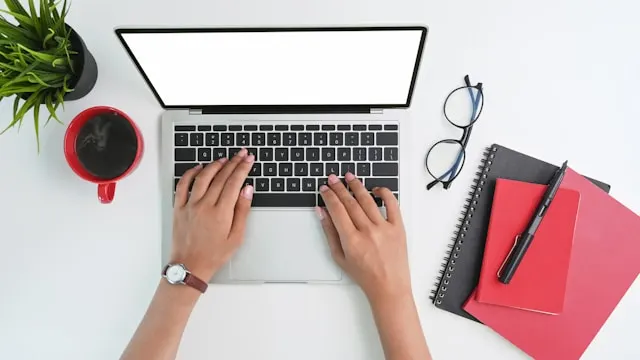 Hands typing on a laptop keyboard, which is on a table along with a cup, glasses, notebooks and pen.