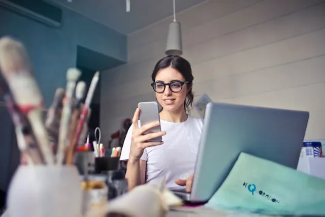 A young woman with glasses working on her laptop and looking at her phone.