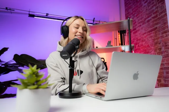 A woman with headphones speaking to a microphone while using a laptop.