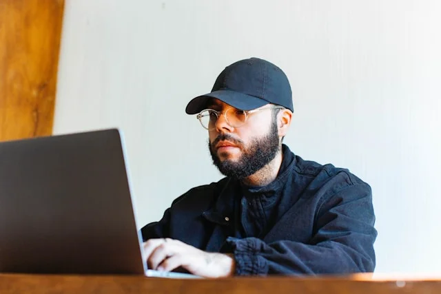 A man with a cap and glasses using a laptop.