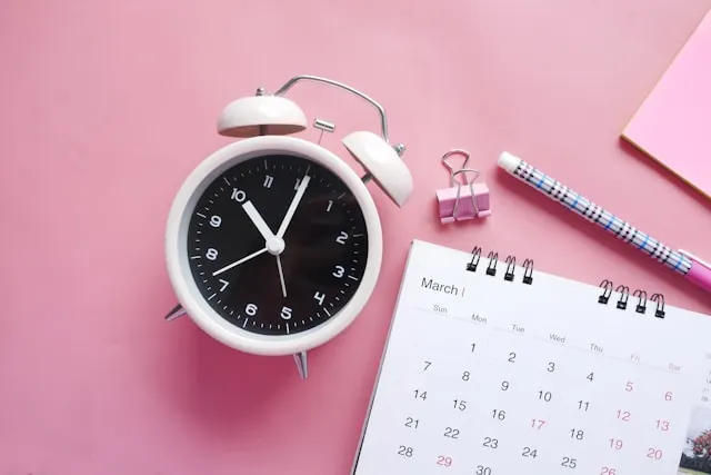 A calendar and and alarm clock on pink surface.