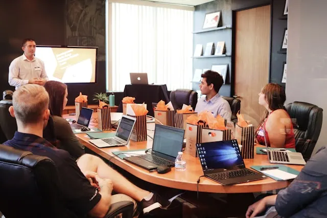 Man standing in front of people who are sitting around a table and watching the presentation.