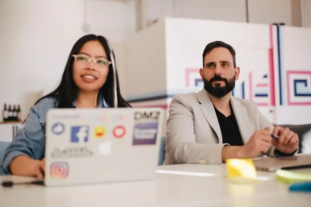 Two people in a meeting, seated at a table with laptops open in front of them.