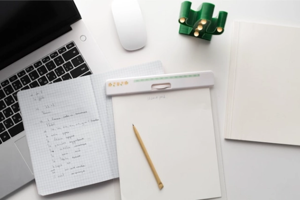 A laptop, a notebook, a pen, and sheets of paper on a desk.